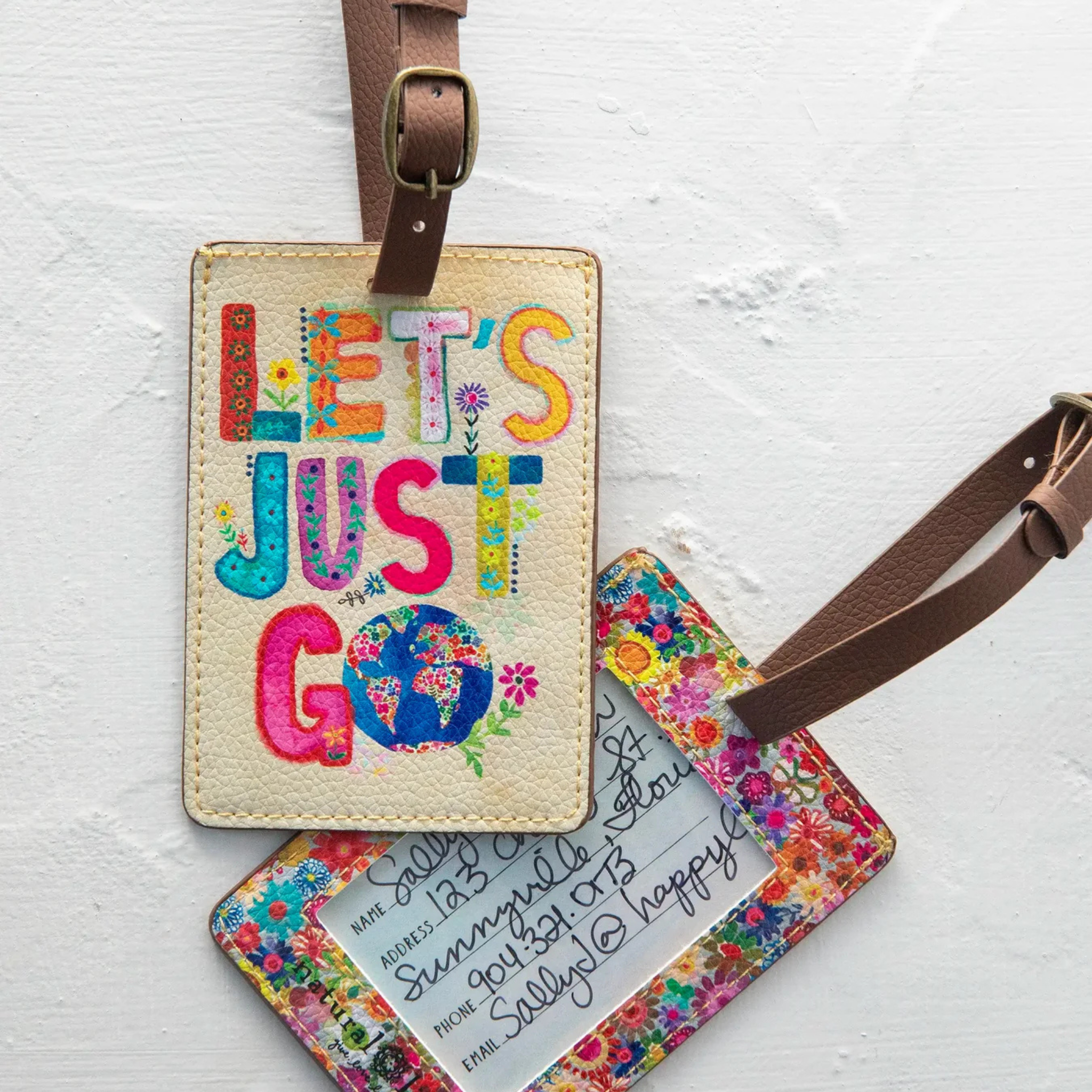 Natural Life Travel Happy luggage tag with "Let's Just Go" message, featuring colorful design and durable strap for travel convenience