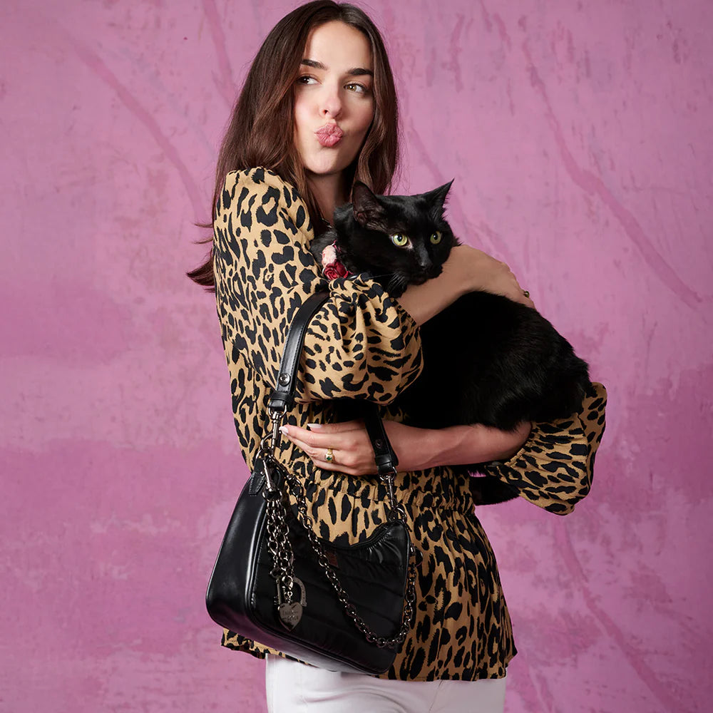 Woman in a leopard print top holding a black cat, showcasing a Think Royln Soulmate shoulder bag against a pink background.